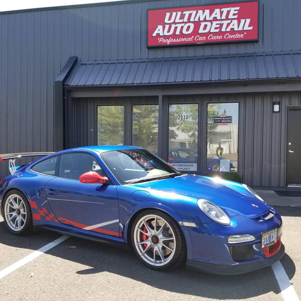 A blue porsche with a red checker stripe parked in front of the ultimate auto detail shop